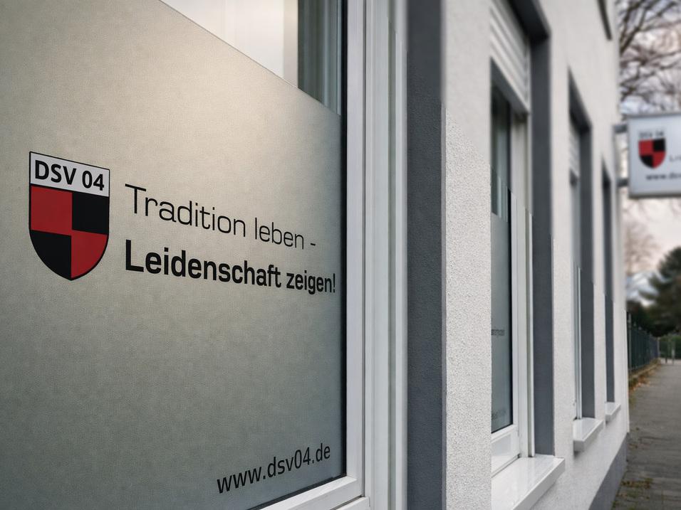 Fenster mit Schriftzug „Tradition leben - Leidenschaft zeigen!“ an einem hellen Gebäude, Straßenszene im Hintergrund.