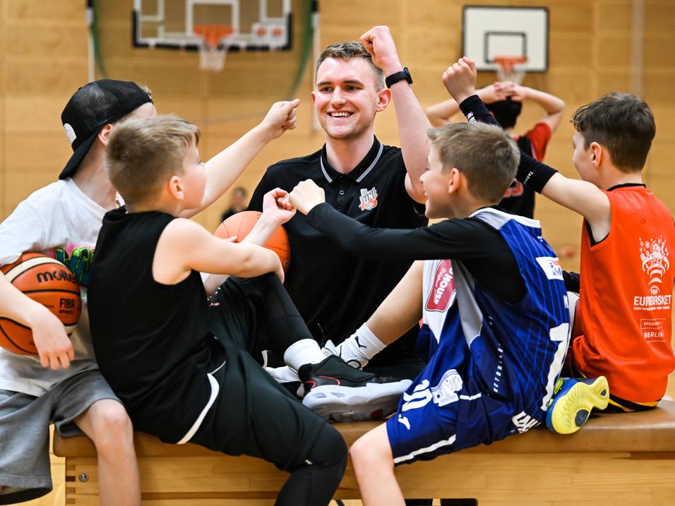 Sechs Kinder und ein Trainer zeigen Freude beim Basketballtraining auf einer Holzbank in einer Sporthalle.