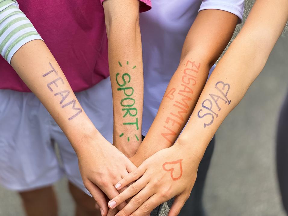 Vier Arme liegen übereinander, mit farbiger Schrift: „TEAM“, „SPORT“, „ZUSAMMEN“, „SPASS“ auf der Haut.